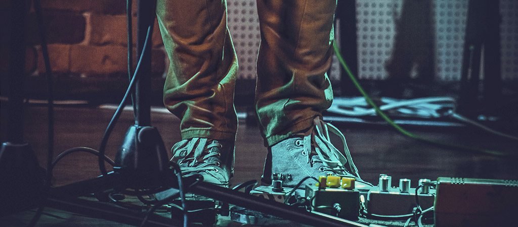 A closeup of the feet of a person near guitar pedals and a mic stand under the lights