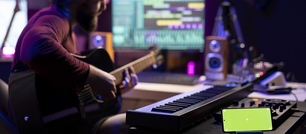 Young singer playing guitar and practicing with online tutorial lessons, looks at daw software on pc. Musician learning new acoustic instrument accords, having phone with greenscreen display.